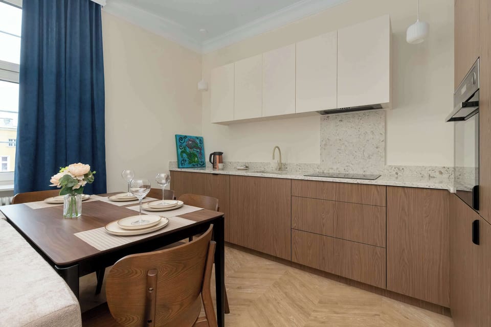 An elegant dining and kitchenette space includes a dark wooden table and a streamlined kitchen with wood-textured lower cabinets and white upper units. The herringbone parquet flooring adds a classic touch to the modern interior.