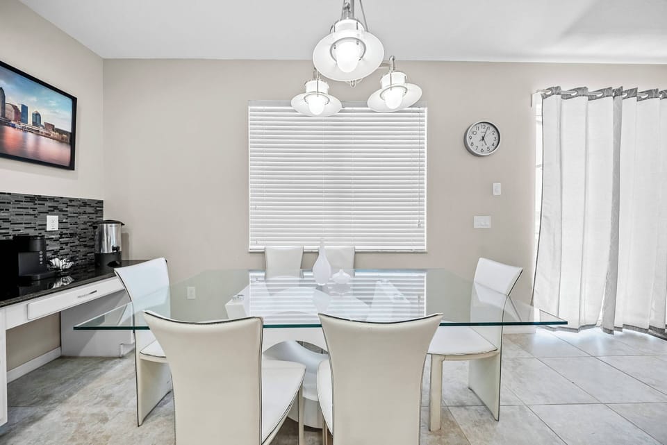 This dining area features a sleek glass table surrounded by modern white chairs, creating a bright and airy atmosphere. Soft, natural light pours in through the adjacent window, enhancing the subtle elegance of the space.
