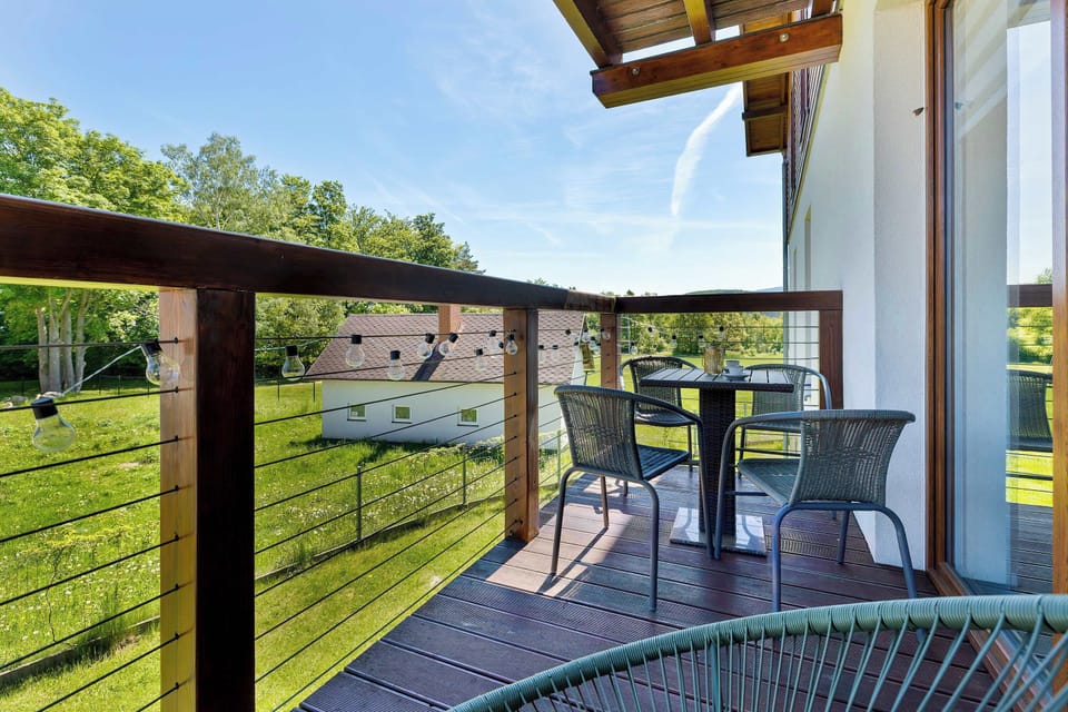 Another view of the terrace or balcony overlooking the neighboring Alpine-style building and the surrounding greenery. A set of outdoor furniture consisting of a table and chairs is placed by the railing.