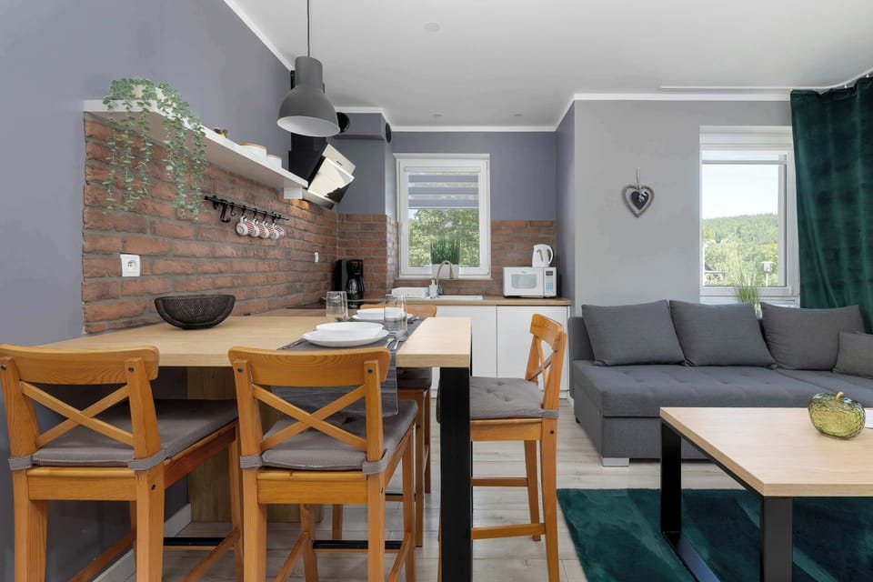 The dining area features a large wooden table and matching chairs, illuminated by industrial-style pendant lights. In the background, a section of the gray sofa and a window overlooking greenery are visible.