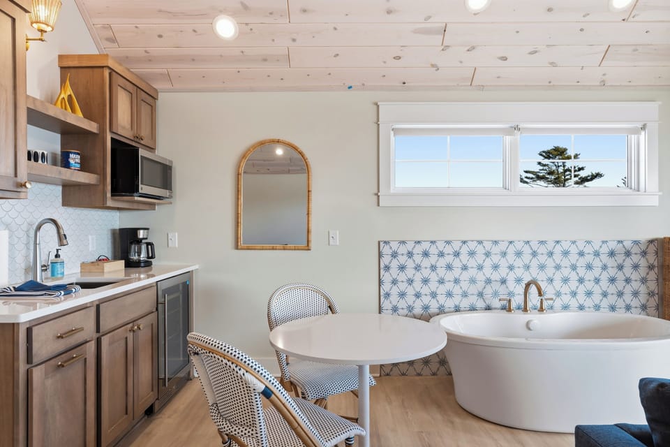 Step into your charming coastal retreat, where a cozy kitchenette and a dreamy soaking tub meet under white pine ceilings, with glimpses of the shoreline through the window.