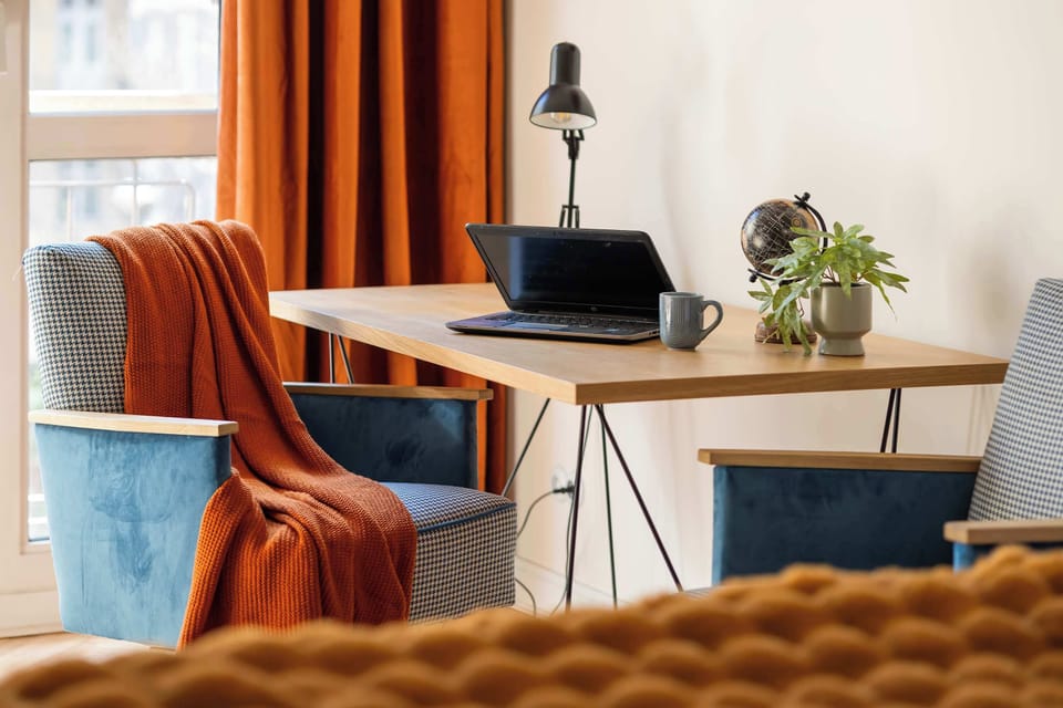 A cozy corner features a desk by a window with sheer curtains, a blue velvet chair with a warm orange throw blanket, a laptop and lamp on the desk, and a large orange throw in the foreground.