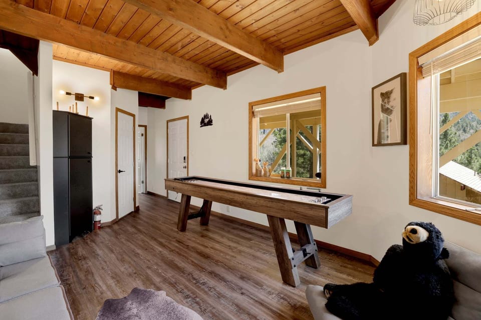 A cabin-style game room with a shuffleboard table, a gray sofa, exposed wood ceiling beams, and dual TV screens.