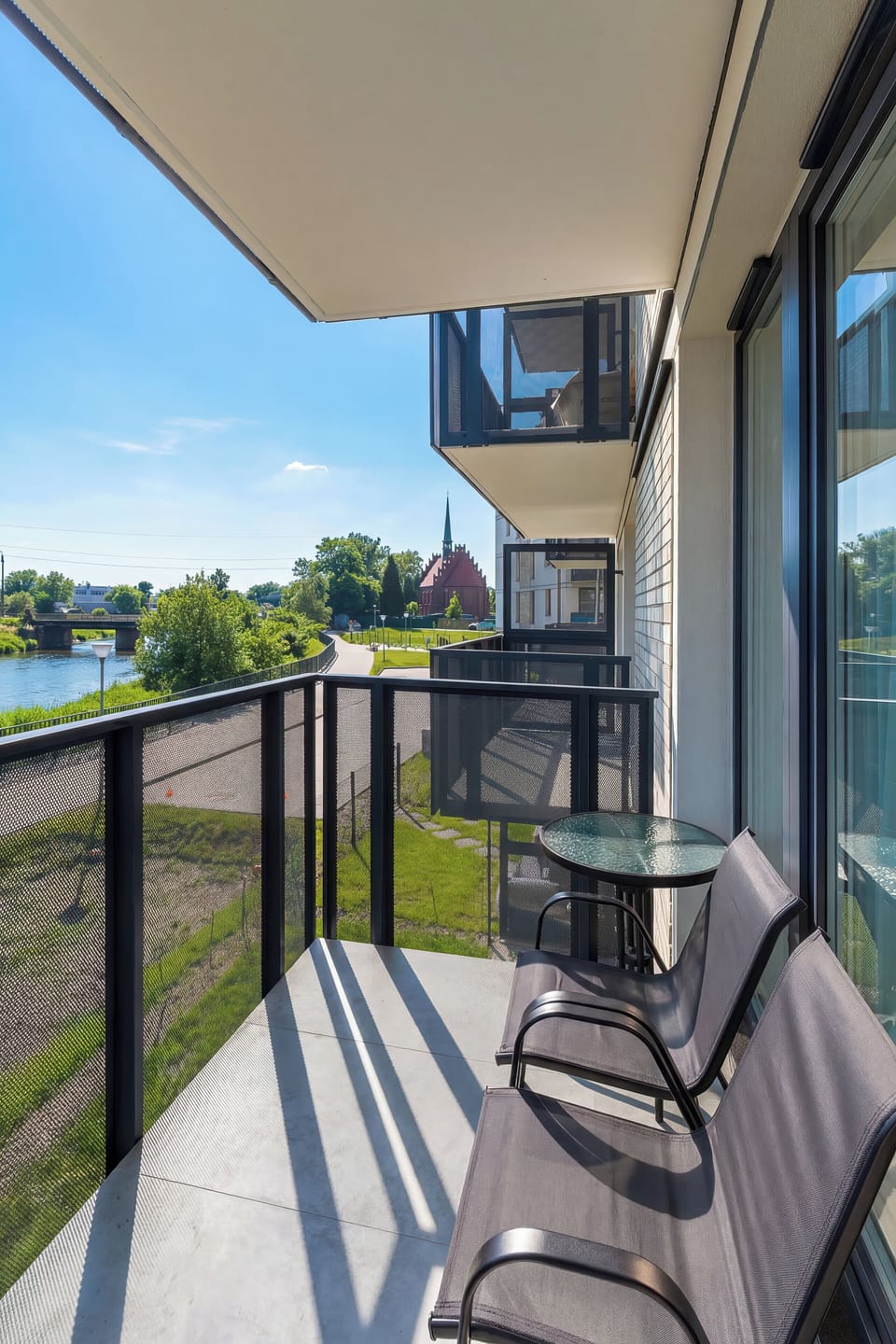 From a high-angle perspective, the balcony shows off its glass railing and two comfortable outdoor chairs. The view extends toward a quiet street and green landscape under a clear blue sky.