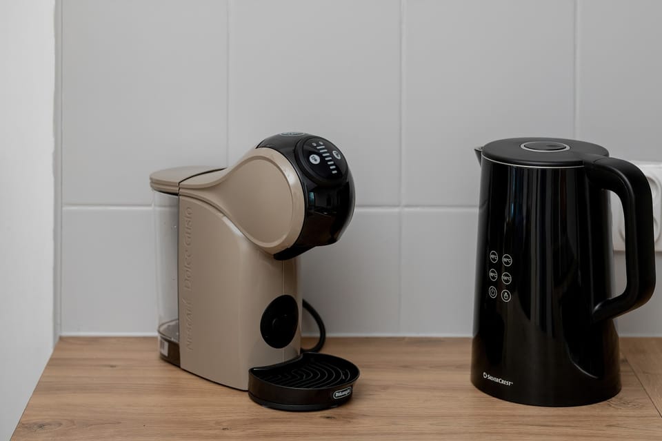 Close-up of a coffee station with a capsule coffee machine and an electric kettle. The setup sits on a wooden countertop against a white wall.