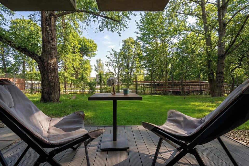 An outdoor terrace is furnished with two modern chairs and a small table, ideal for morning coffee. The space overlooks a lush green garden area with mature trees.