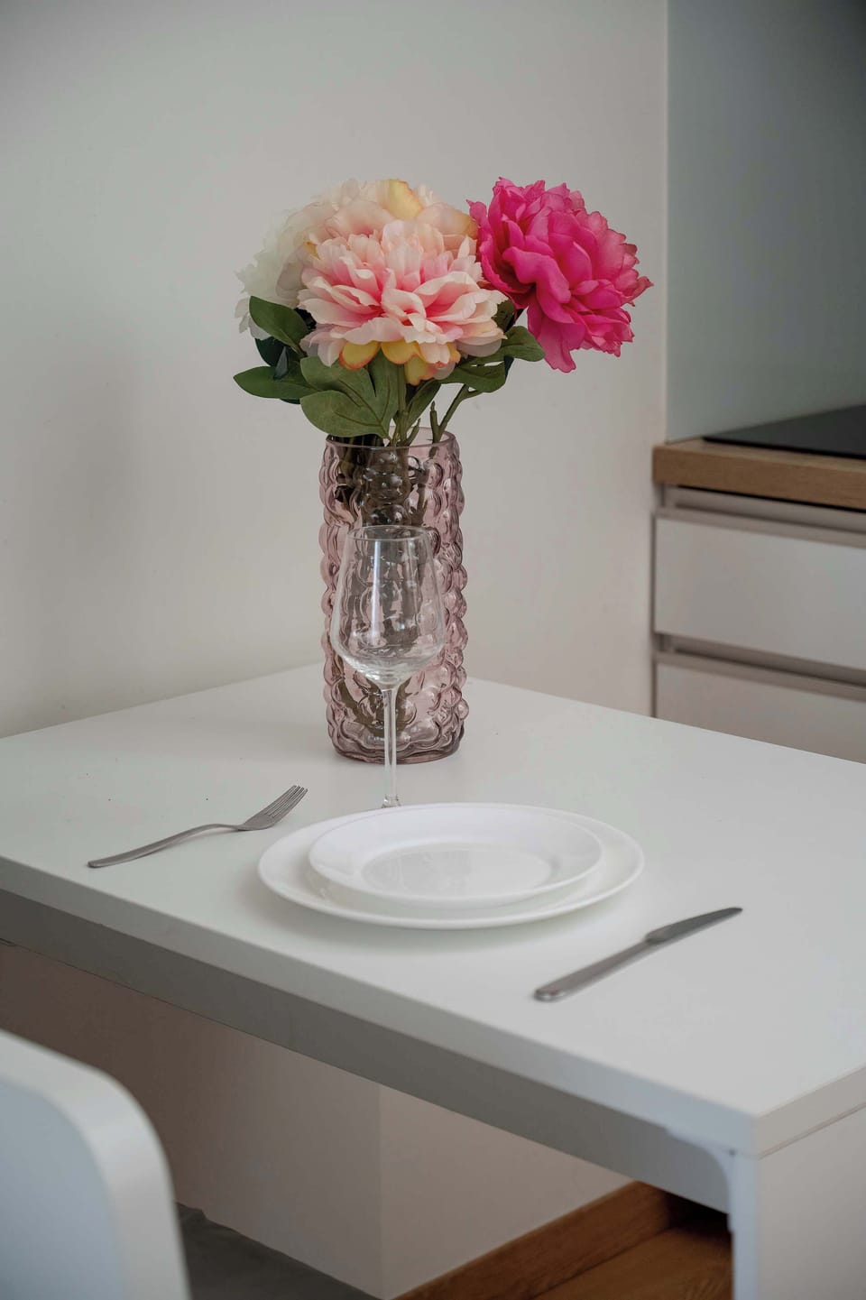 A detailed shot of the dining table focuses on a tall glass vase with vibrant pink flowers. The setting is simple and clean, matching the minimalist aesthetic of the entire apartment.