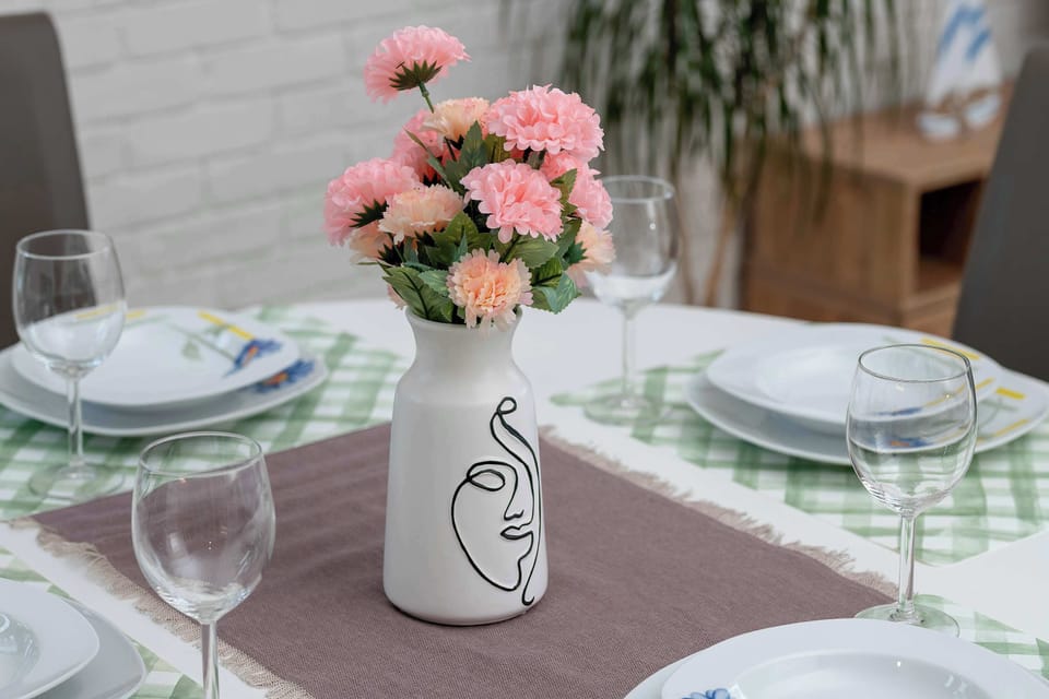 A round white dining table is elegantly set for four people with white plates and glassware. A stylish vase with pink flowers serves as a centerpiece against the backdrop of a white brick wall.