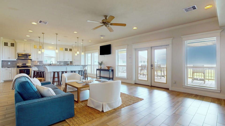 Bright open-concept living room with coastal décor, large sectional seating, ceiling fans, and wall-to-wall windows with beach views.