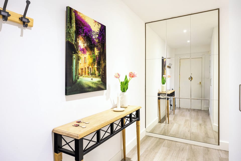 A bedroom, a desk, and air conditioning. Bedside tables with a lamp, a wooden dresser, window, a desk with a chair and a lamp on it. A mirror hangs on the wall.