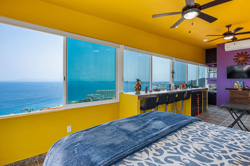 Perspective from the bed overlooking a long window bar with stools facing the blue sea.