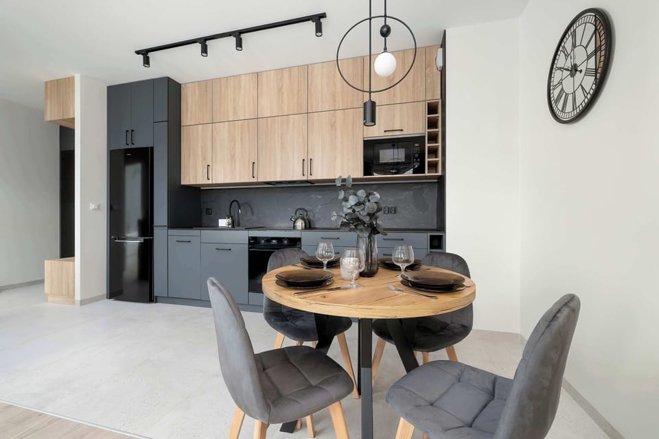 A well-lit kitchen and dining space that balances dark grey finishes with light wood textures and a modern wall clock. The integration of colors creates a sophisticated atmosphere.