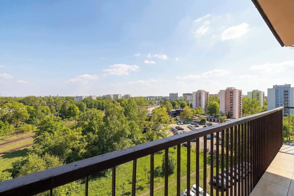 A wide shot from the balcony with a metal railing, offering a soothing view of the treetops and city architecture in the distance.