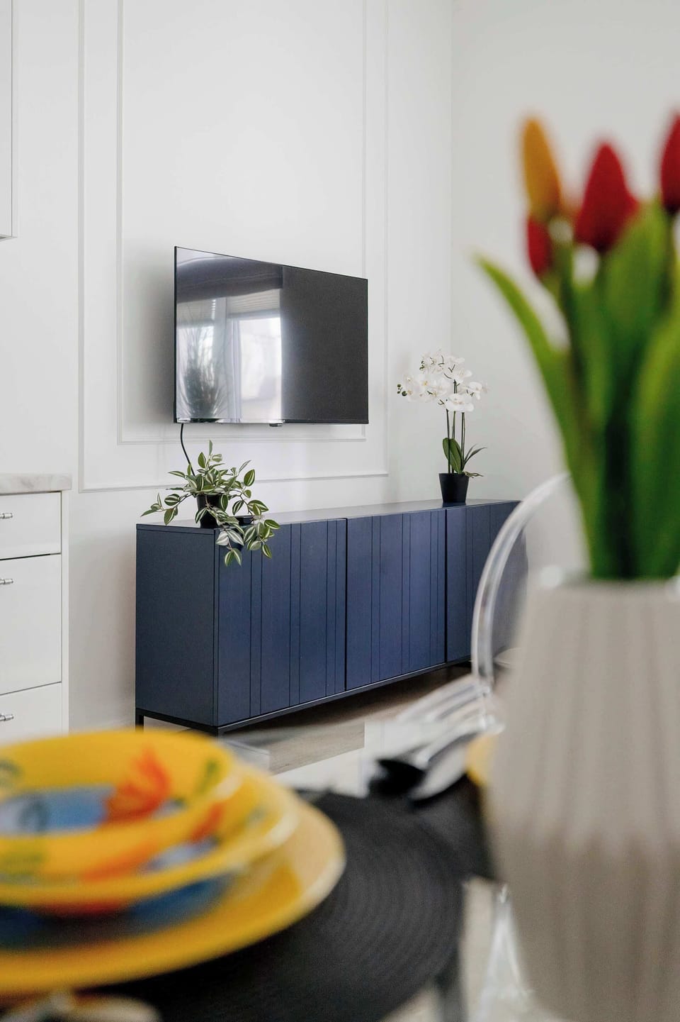 A tightly composed vertical shot focusing on a white nightstand holding a small vase of orange tulips. The dark blue media console and a TV are visible in the background.