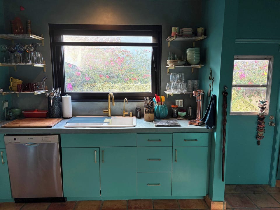 Prep and bar area with teal cabinetry, brass faucet, dishwasher, and open glassware shelving.
