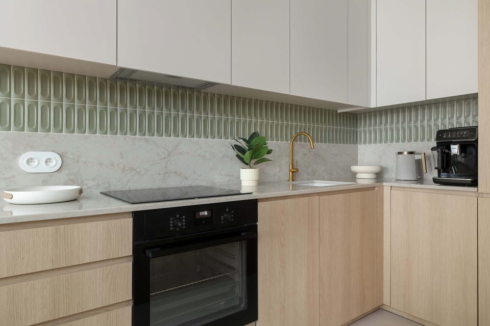 A contemporary kitchenette showcases light wood lower cabinets paired with minimalist white upper units and a green-tiled backsplash. The space is equipped with modern built-in appliances and gold-toned fixtures.