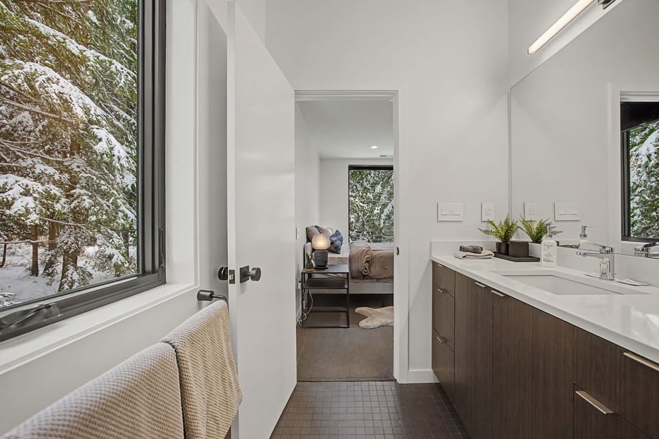 Your private spa-like bathroom features dual vanities and stunning winter forest views through floor-to-ceiling windows.