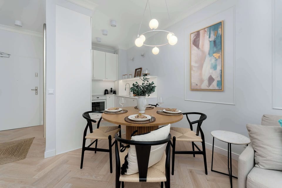 A dining area features a round wooden table with four modern black-frame chairs. A distinctive circular chandelier hangs above the table, and an abstract artwork decorates the wall. The space is adjacent to the open-plan kitchen and a doorway.