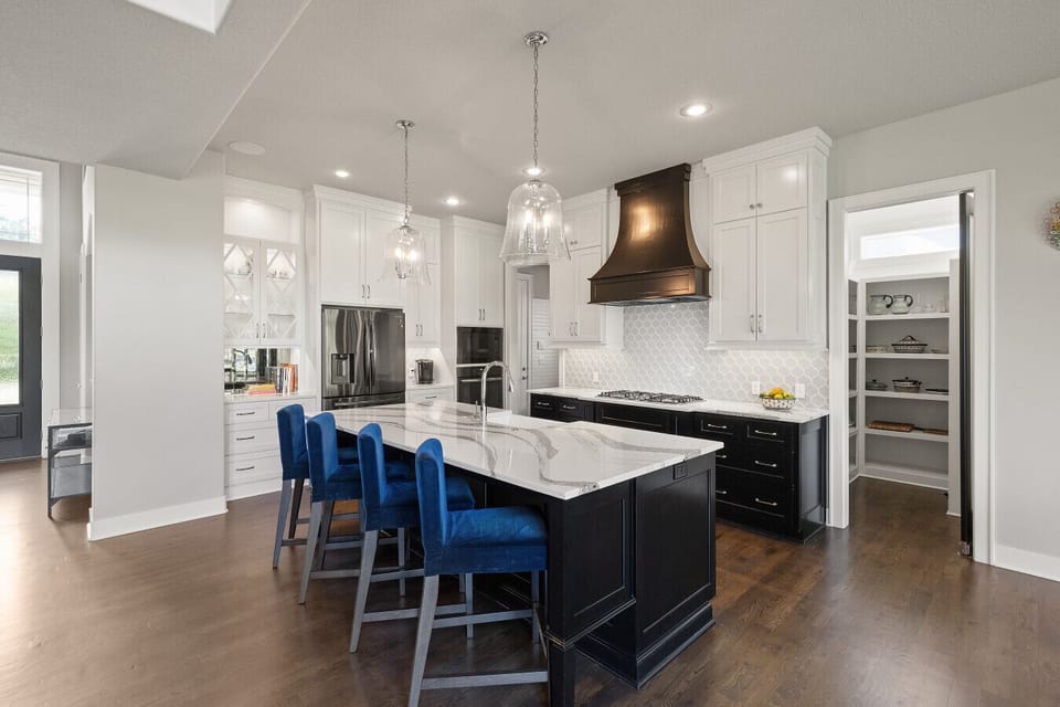 The heart of this home is a bright, luxurious kitchen with sleek black cabinets, sparkling quartz countertops, and vibrant blue barstools waiting for a lively gathering.
