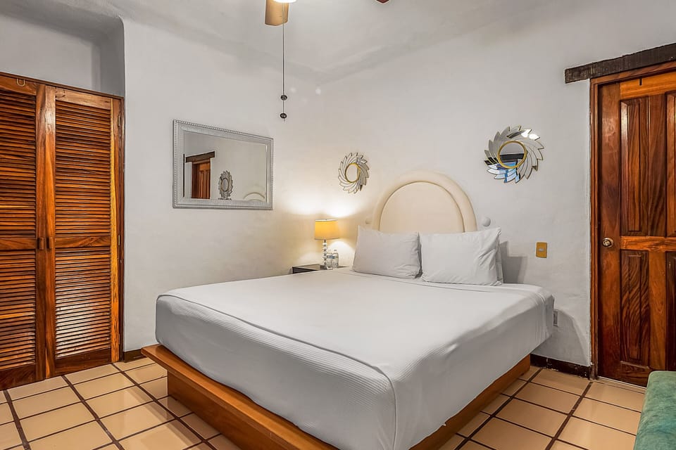 Comfortable master bedroom featuring a king-sized platform bed with crisp white linens, a large rectangular mirror alongside decorative accents, and traditional louvered wood closets for guest storage.