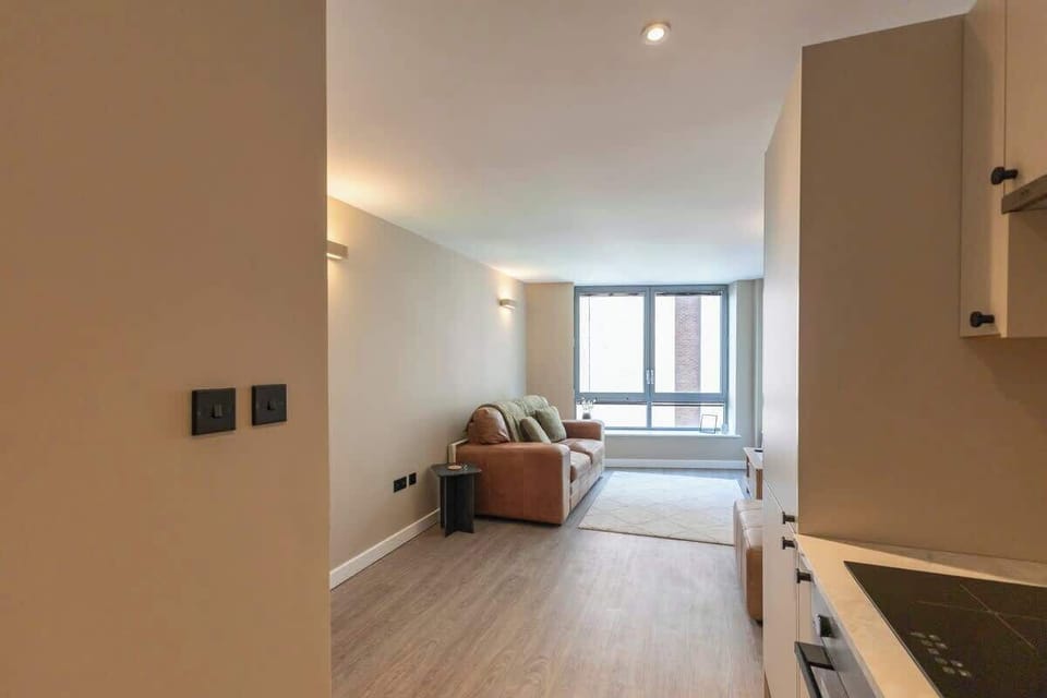 Bright and modern living room with large windows that fill the space with natural light and city views. Features a cozy leather sofa, sleek wooden accents, and a minimalist TV setup for a relaxing stay.