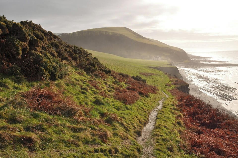 Coastal footpath along grassy cliff with waves and distant hills