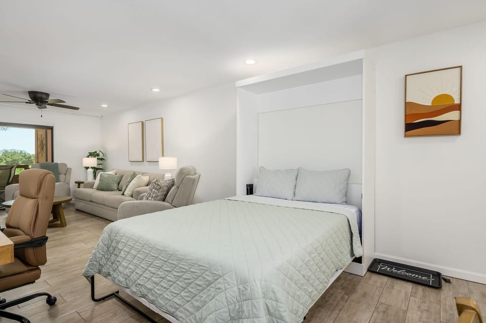 Murphy bed with striped pillows folds out beside a tan office chair and beige sofa in the living area.
