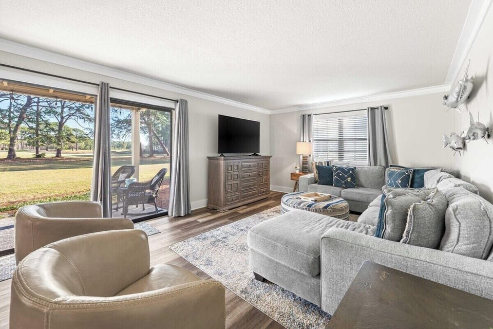 Sunlit living room with oversized sectional, flat-screen TV, and sliding doors opening to the greens — lounging has never been this easy