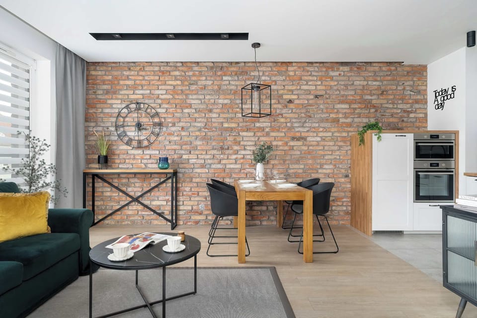 The exposed brick wall serves as a texture-rich backdrop for a rustic wooden dining table and black-seated chairs. Two contemporary pendant lights with geometric shades illuminate the dining area, enhancing the modern feel.