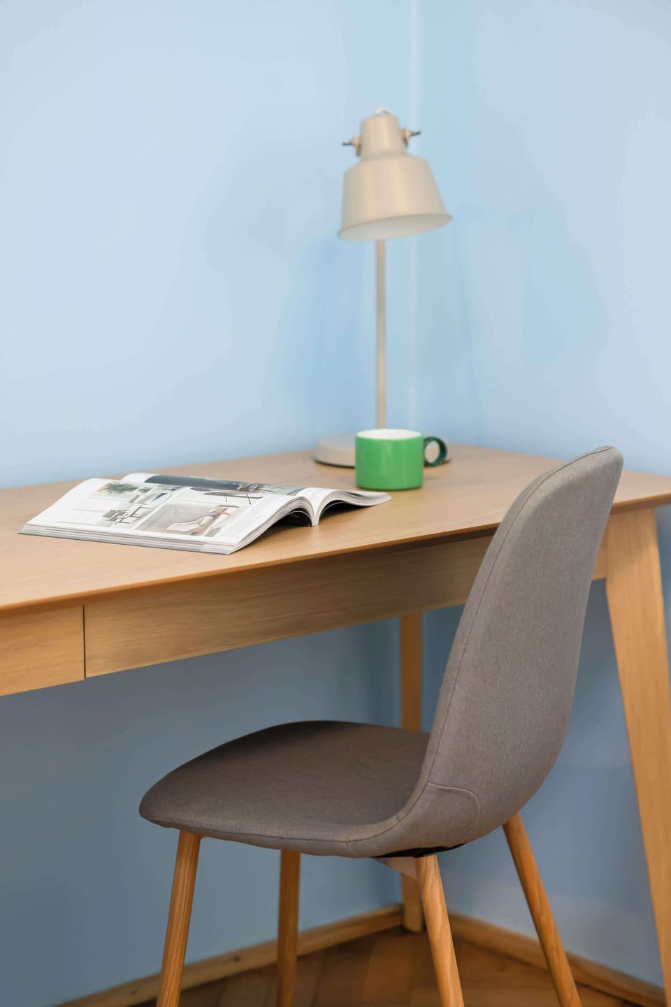 A minimal study nook containing a simple wooden desk, a grey-upholstered chair, and a unique, slender desk lamp, all against a clean blue wall.