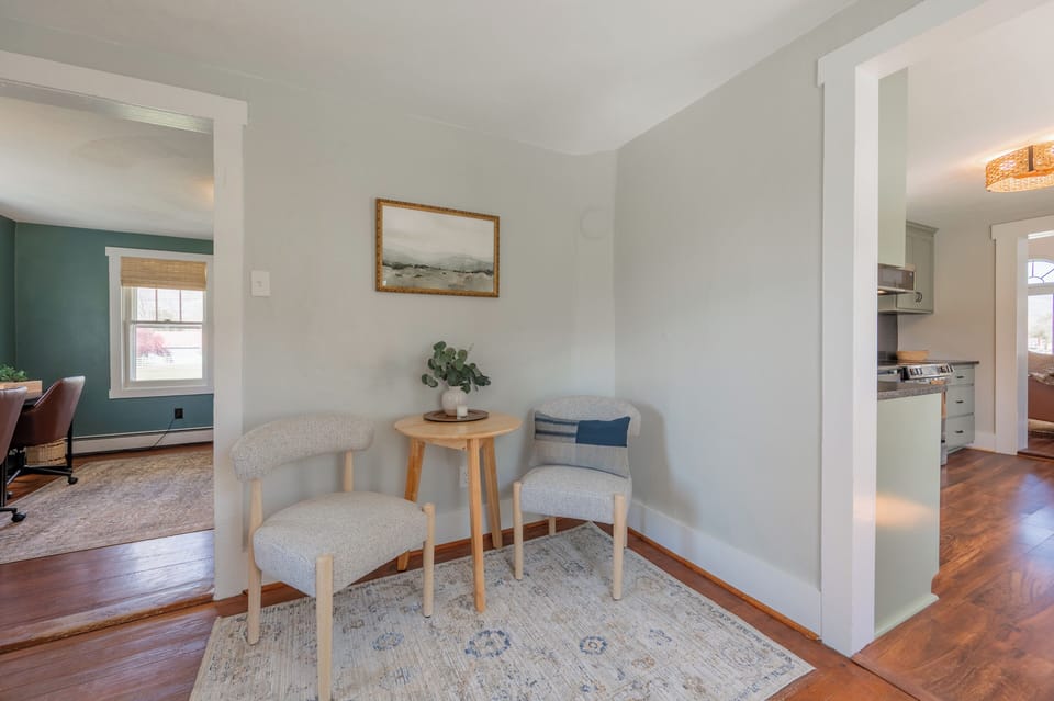 A charming seating nook featuring two upholstered chairs, a small wooden bistro table, and a patterned area rug, perfectly positioned between light-filled rooms.