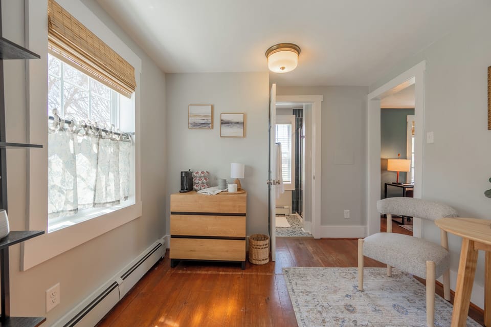 A bright, neutrally decorated room featuring light wood flooring, a coffee station on a wooden dresser, and a cozy seating area.