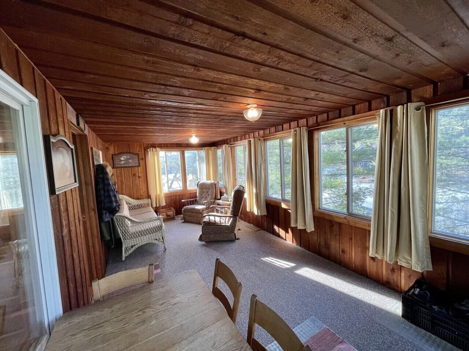 Unwind in your rustic sunroom with warm wood walls and panoramic windows showcasing peaceful lake views.