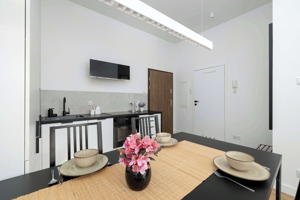 A wooden dining table is set with dark placemats and ceramic bowls, ready for a meal. A vase with pink flowers sits in the center, adding a pop of color to the monochrome kitchen background. 