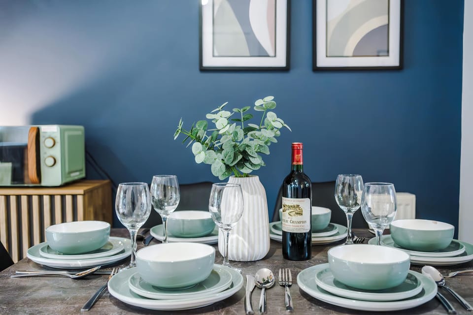 Stylish dining area with a set table, wine centerpiece, and greenery. The deep blue wall, framed art, and staircase detail add sophistication, blending warmth and elegance for a guest‑friendly dining experience.