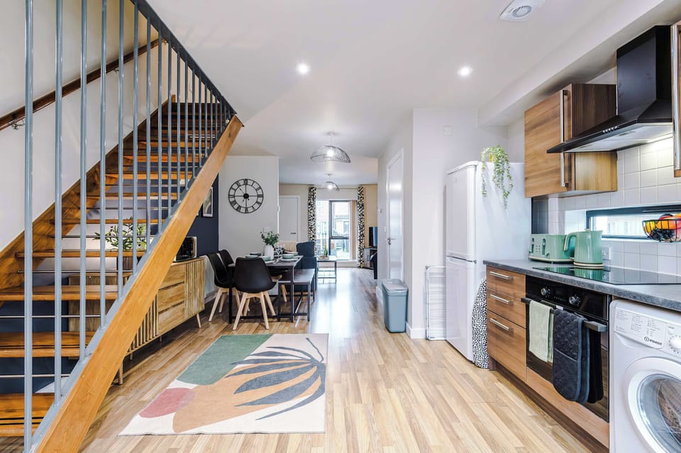 Bright, open living space with a geometric rug, sleek staircase, and cozy dining area. Decorative accents and framed art enhance the ambiance, blending clean lines with warmth for a guest‑friendly, contemporary atmosphere.