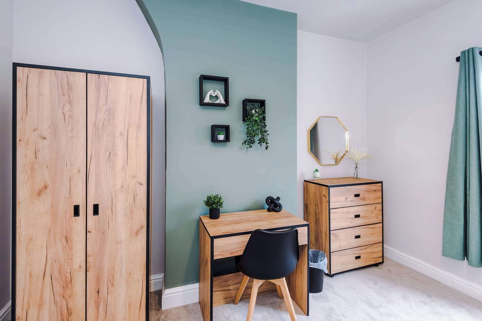 Contemporary comfort: a spacious bed with white bedding and patterned accents, warm-lit nightstands, framed art on a muted green wall, and large windows with green curtains letting in natural light.