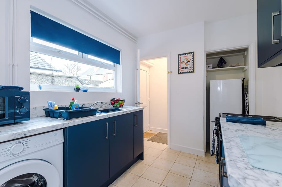  A bright, modern kitchen with blue cabinetry, white marble counters, and a washing machine built-in. A coffee machine, fruit bowl, and dish rack add charm, while natural light from the window enhances the clean, stylish design.