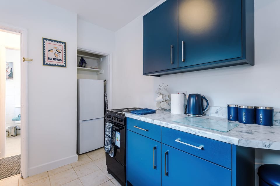 Contemporary kitchen design featuring blue cabinets, marble countertops, and practical appliances. A dish rack, coffee machine, and fruit bowl bring everyday warmth to the organized, light-filled space