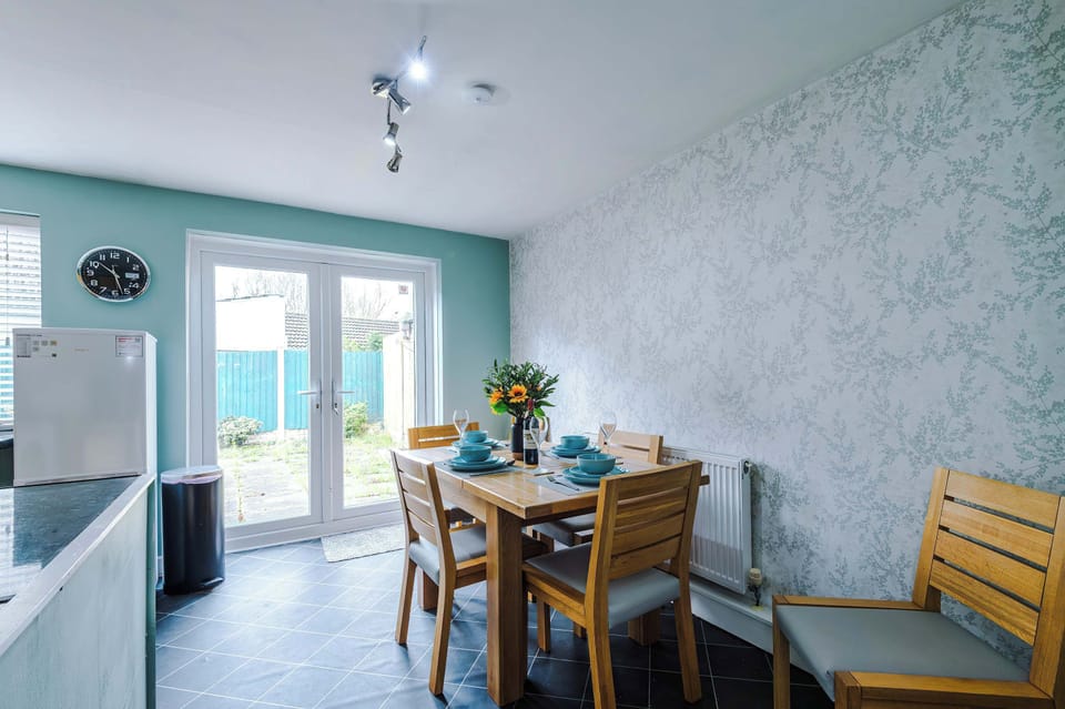 A modern dining area set with wine glasses, blue plates, and sunflowers, opening into a cozy living room. The textured wall and dark tile floors add depth to the bright, inviting space.
