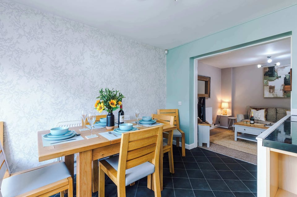 A warm dining space with a wooden table set for four, blue plates, wine glasses, and sunflowers as a centerpiece. Through the doorway, a cozy living room with sofa and shelves completes the inviting home design.