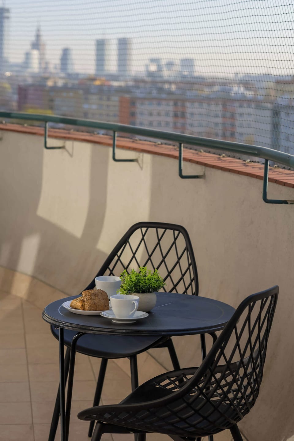 A balcony features a small table with coffee, croissants, and a small plant. The safety netting allows for clear views of the distant city skyscrapers and rooftops.
