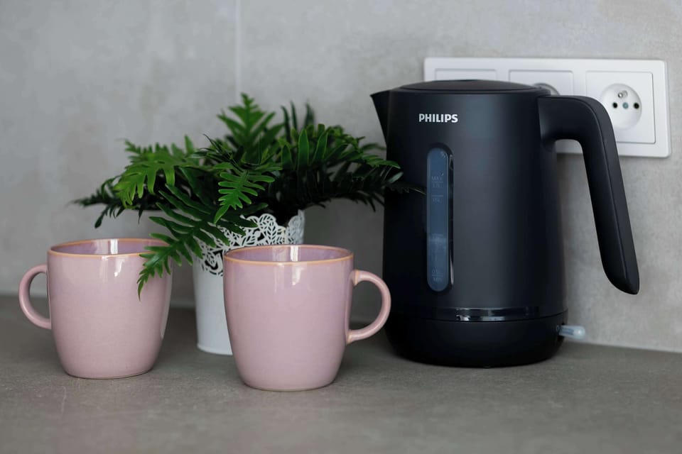 A black electric kettle stands on the kitchen counter next to two matte pink mugs and a small green plant. This lifestyle shot highlights the functional and aesthetic details of the kitchen.