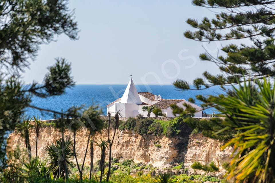 Stunning panoramic sea view from 813 Alporchinhos, looking across dramatic golden cliffs and a charming white chapel towards the sparkling Atlantic Ocean, framed by pine trees and Mediterranean vegetation in the Algarve