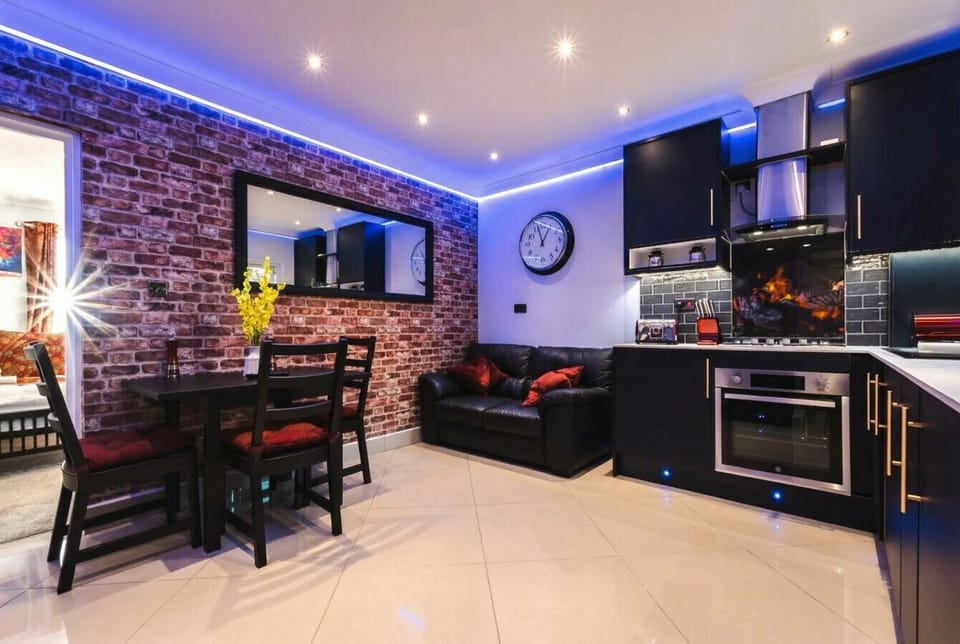 The open-plan living space glows under blue LED ceiling lights, where exposed brick, a leather sofa, dining table, and fitted kitchen all flow together.