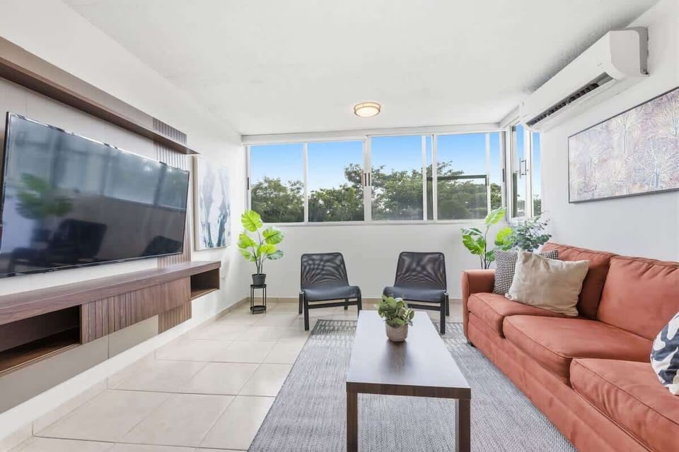 Bright, modern living room with large windows, a wall-mounted TV, and a warm terracotta sofa.