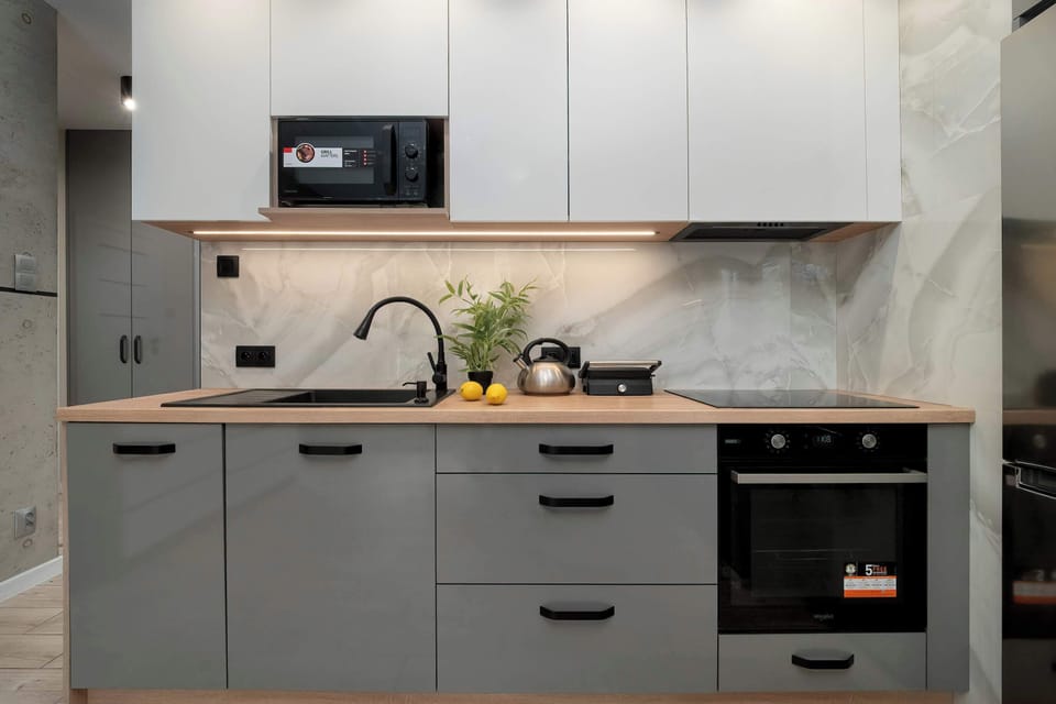 A compact, modern kitchen workspace is equipped with grey cabinetry and a light wood-toned countertop. An oven and a microwave are integrated into the unit for a seamless look.