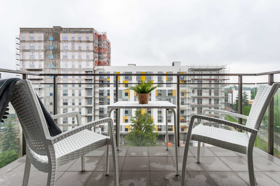 A private balcony is furnished with two white chairs and a small table, overlooking a neighboring modern building. A small yellow plant adds a splash of color to the outdoor space.