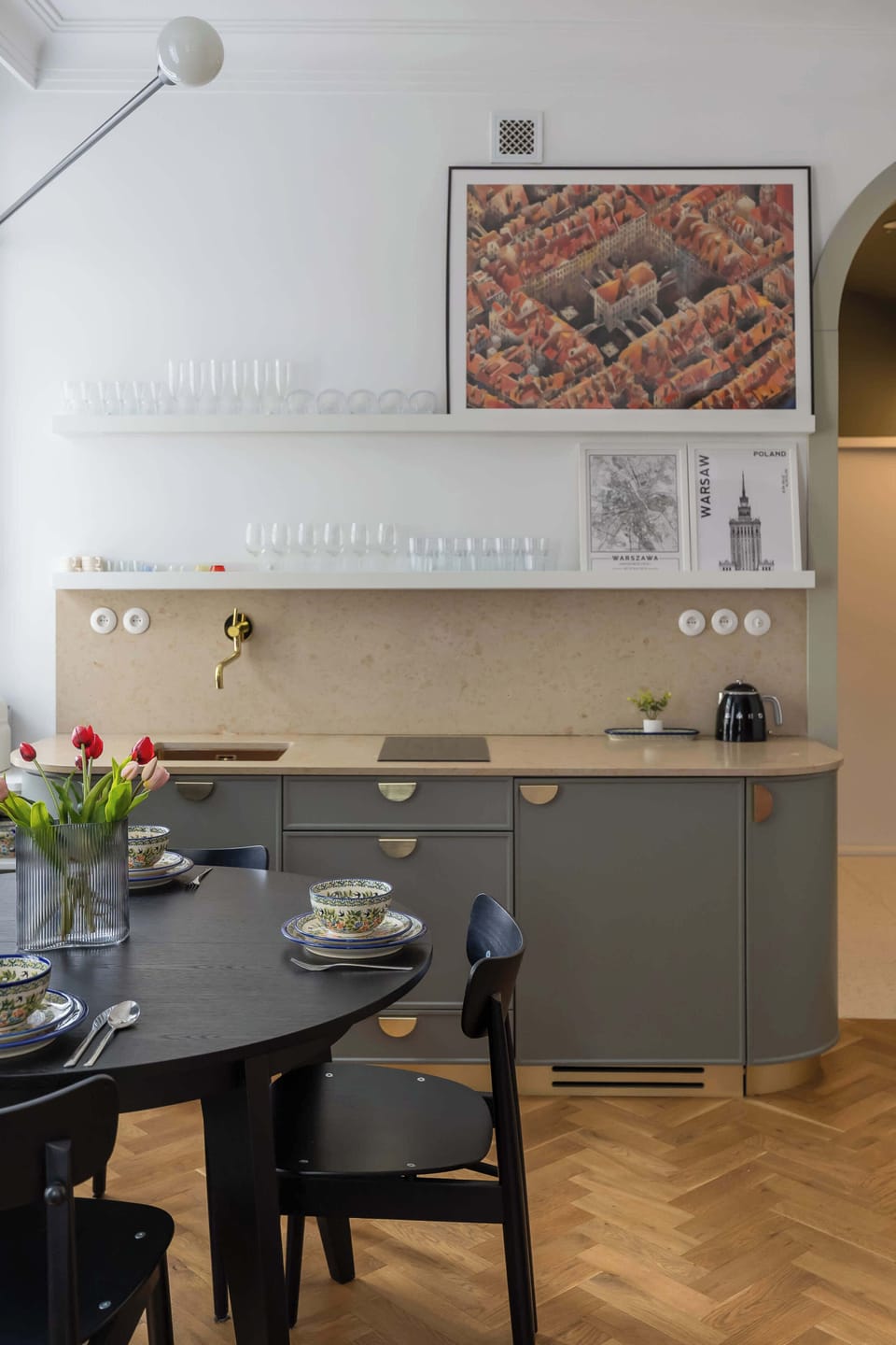 A bright, high-ceiling kitchen area contains grey cabinets and black dining chairs surrounding a dark table. A decorative, textured textile hanging adds a touch of art to the neutral wall.