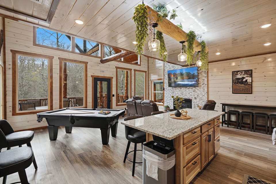 Unwind in your spacious game room with pool table, cozy fireplace, and stunning forest views through floor-to-ceiling windows.
